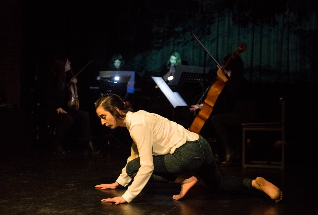 A dancer on the floor in the foreground; violin, voice and cello playing behind.