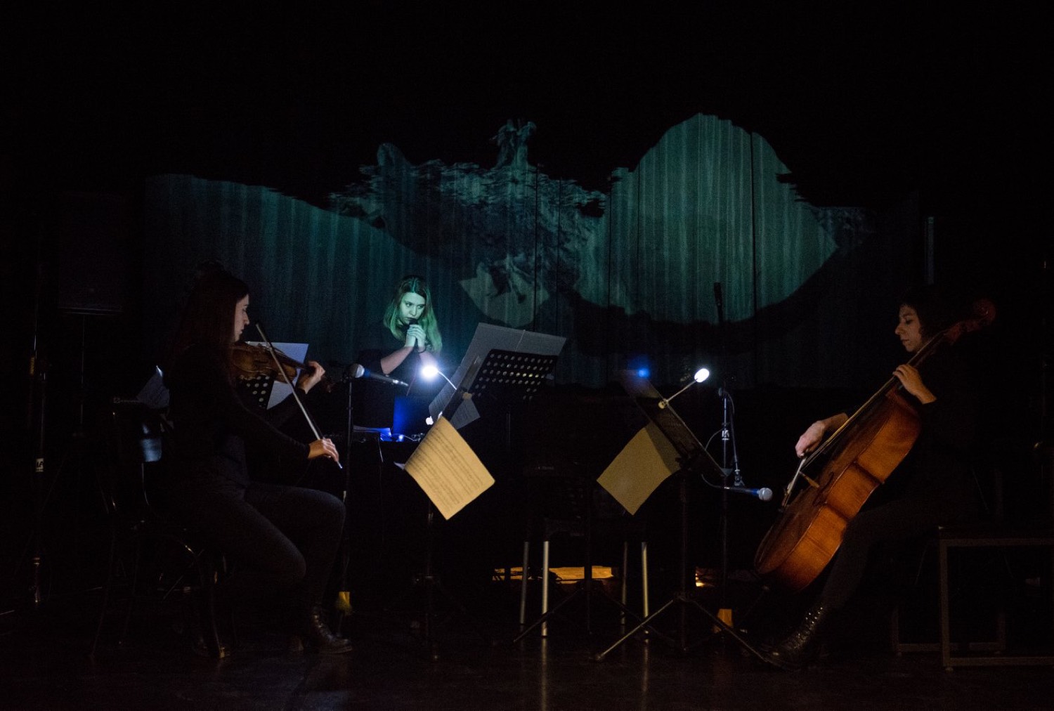 klank.ist in performance: violin, voice and electronics, and cello in front of a projection.