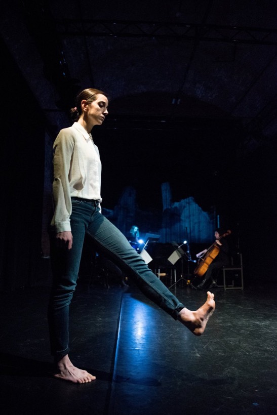 A dancer mid-gesture in a dark hall, with violin and cello playing behind a blue projection.