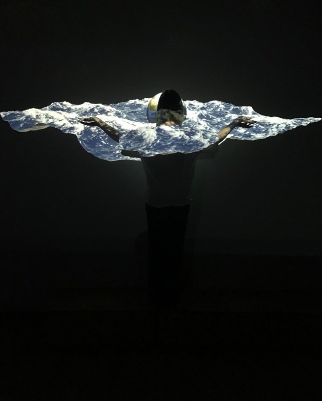 A performer with arms outstretched, a projection of a sea breaking across their body in a dark room.